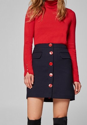 Femme portant un col roulé rouge à manches longues et une jupe bleu marine avec de gros boutons rouges sur le devant.