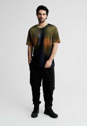 Mężczyzna o ciemnych włosach stoi, ubrany w zielono-pomarańczowy t-shirt z gradientem, czarne spodnie typu cargo i czarne buty na jednolitym jasnym tle.