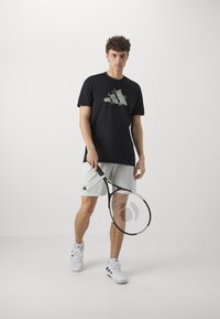 T-shirt noir à manches courtes avec un design graphique, associé à un short gris et des chaussures de sport blanches, tenant une raquette de tennis.