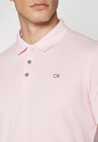 Šviesiai rožinė polo marškinėliai su trumpomis rankovėmis, apykakle ir trimis pilkais mygtukais. Kairėje krūtinės pusėje išsiuvinėtas "CK" logotipas. Tekstūruota medžiaga.