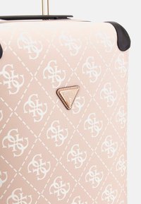 Valise rigide en matériau texturé rose avec un motif de logo blanc. Comprend une plaque de logo triangulaire dorée et des accents noirs.