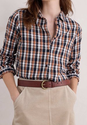 Frau trägt ein langärmeliges kariertes Hemd in Orange, Navy und Weiß, in beige Kordhosen mit einem braunen Gürtel gesteckt, Hände in den Taschen.