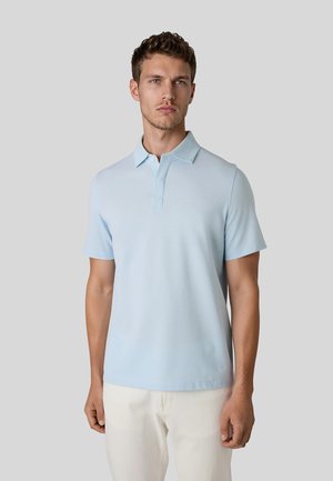 Ung mand med kort krøllet hår iført en lyseblå polo t-shirt med korte ærmer og off-white bukser, stående mod en ensfarvet grå baggrund.