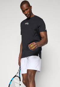 Mann in schwarzem Asics-Tennisshirt und weißen Shorts hält Tennisball und Schläger und blickt auf den Ball hinunter.