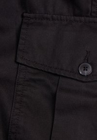 Svart tyg med texturerad yta, utrustad med knappstängning och en fickklaff. Innehåller sydda detaljer längs kanterna.