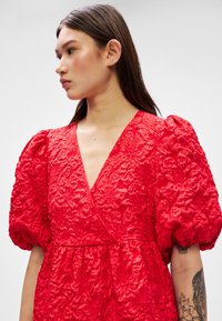 Raudonos suknelės su pūkuotomis rankovėmis, gėlių rašto audinys, V formos iškirpė, priglustanti juosmena. Suknelė turi plazdantį sijoną ir išsamius puošybos elementus.