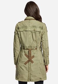 Olivgrön trenchcoat med bälte i midjan, axelklaffar och tryckknapp detaljer. Har ett texturerat tyg och ett korsdesign bak.