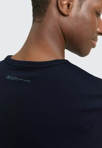 Granatowy bawełniany t-shirt z okrągłym dekoltem. Na plecach znajduje się małe logo marki w jasnoniebieskim kolorze, prezentujące gładką fakturę.