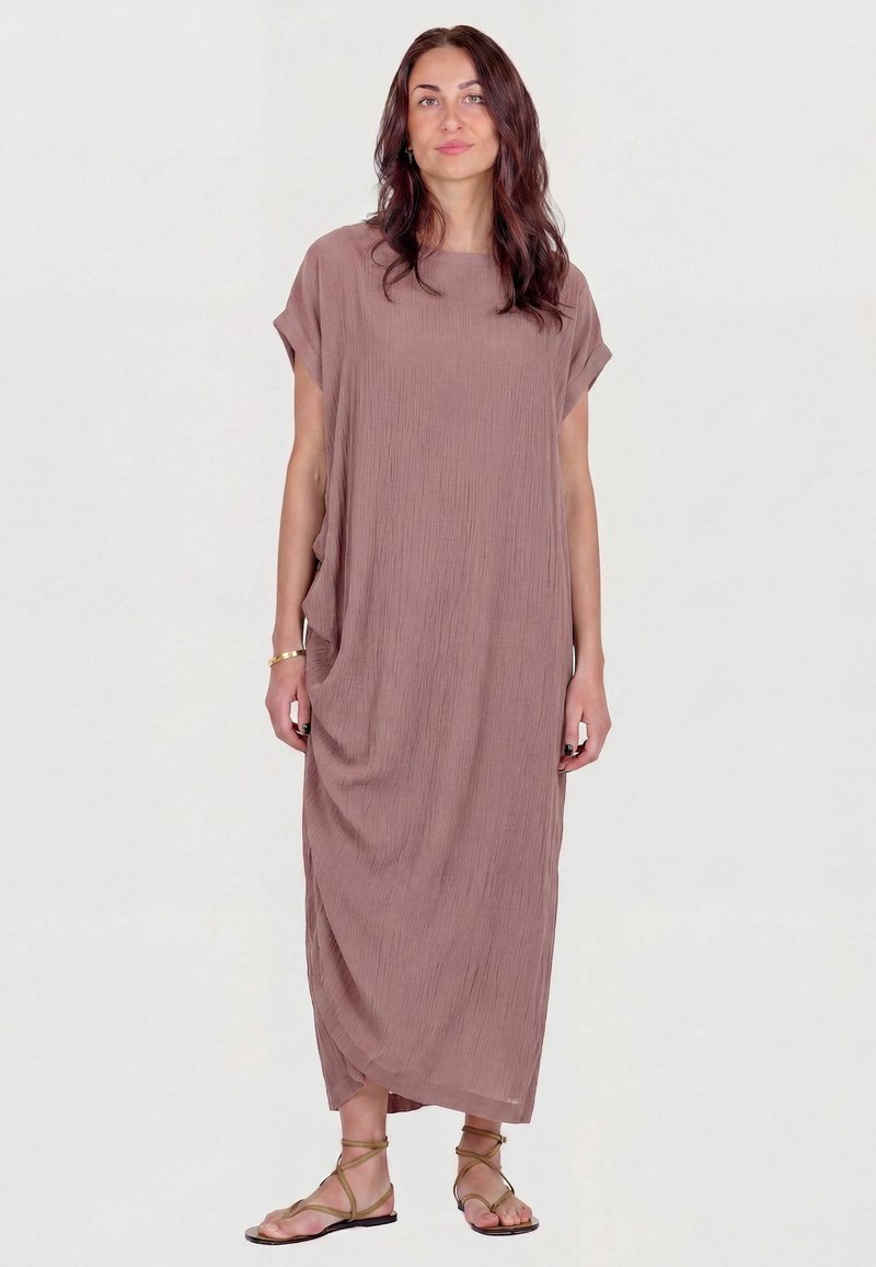 Femme aux longs cheveux bruns portant une robe mauve ample jusqu'aux chevilles et des sandales plates à brides, debout devant un fond uni clair.