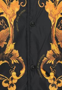 Țesătură neagră cu modele florale baroce galbene și două nasturi negri etichetați "Versace Jeans Couture."