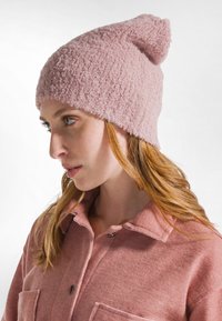 Mujer con cabello largo y rojo que lleva un gorro rosa texturizado y una chaqueta rosa a juego con botones, contra un fondo claro y simple.