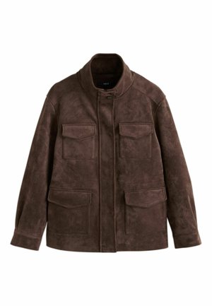 Femme debout devant un fond beige, portant une veste marron, un débardeur blanc, un pantalon large bordeaux, des baskets beiges et tenant un sac à main foncé.