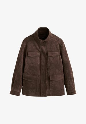 Femme debout devant un fond beige, portant une veste marron, un débardeur blanc, un pantalon large bordeaux, des baskets beiges et tenant un sac à main foncé.