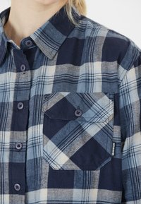 Marineblaues und hellblaues karierte Flanellhemd mit spitzem Kragen, Knopfleiste und Brusttasche. Verfügt über dunkelblaue Knöpfe und ein Etikett an der unteren Seite.