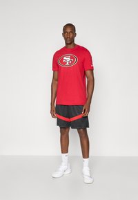 Red short-sleeve t-shirt with SF logo, paired with black shorts featuring red accents. Worn with white athletic shoes and socks.