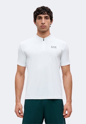 Muž nosiaci biele krátke tričko na zips s logom EA7 Emporio Armani na hrudi a tmavozelené nohavice, stojaci pred jednoliatym pozadím.