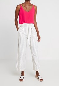 Blouse sans manches fuchsia associée à un pantalon large blanc à rayures verticales, avec ceinture à nouer. Sandales blanches à enfiler complètent la tenue.