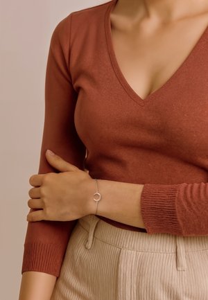 Bracciale d'argento con un design a forma di luna crescentata, indossato su un braccio di pelle chiara. La parte superiore è una maglietta a V di colore ruggine, mentre la parte inferiore presenta strisce verticali color crema.