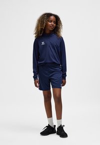 Marineblauw sportoutfit met ritskraag, lange mouwen en bijpassende shorts; gemaakt van lichtgewicht stof; voorzien van logo-accenten en zwarte sneakers.