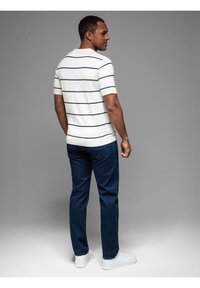 Bílé tričko s krátkým rukávem a námořnickými pruhy, spárované s tmavě modrými džíny a bílými teniskami, viděné zezadu. Hladký materiál, ležérní střih.