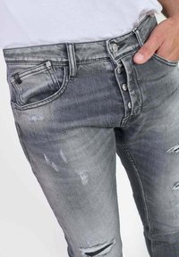 Grijze versleten denim jeans hebben een knoopsluiting, een vijf-pocket ontwerp en een vervaagde was met rafelige gebieden bij de knieën en zakken.