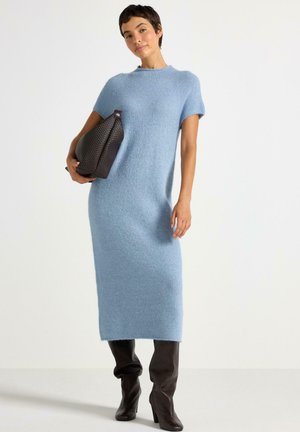 Donna che indossa un vestito midi lavorato a maglia di un azzurro chiaro, tiene una grande borsa intrecciata marrone scuro e indossa stivali fino al ginocchio marrone scuro.