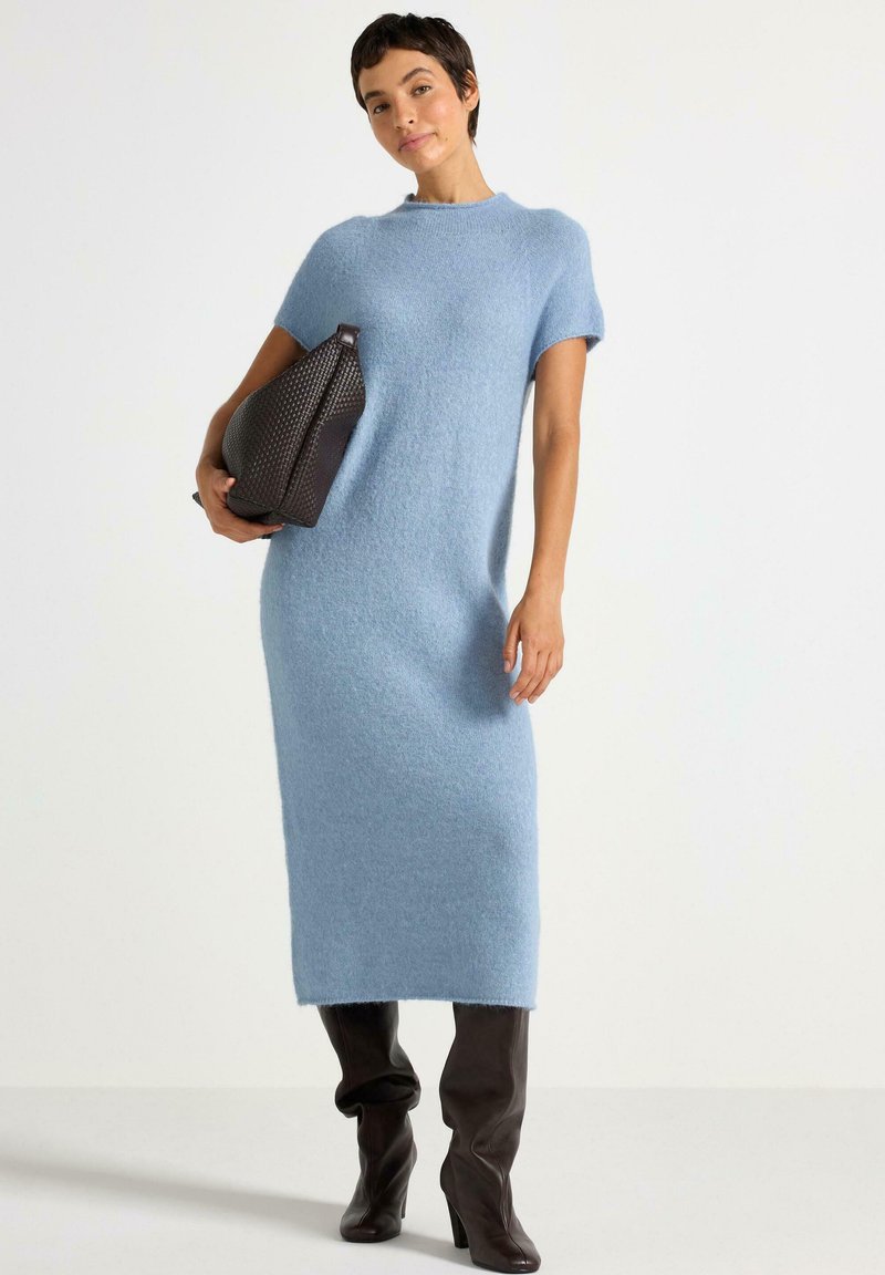 Nainen, jolla on yllään vaaleansininen neule midi-mekko, pitää suurta tummanruskeaa kudottua käsilaukkua ja hänellä on päällään tummanruskeat polvipituiset saappaat.