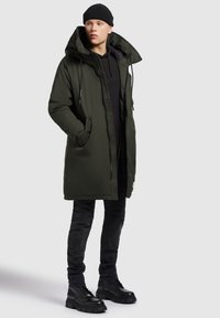 Lang olivengrøn parka-jakke med stor hætte, forreste lynlås og sidelommer, båret over en sort hættetrøje, kombineret med sorte jeans og støvler.