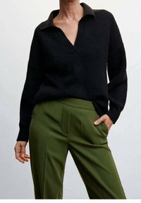 Pull en maille noir avec un col en V, associé à un pantalon vert olive à coupe ajustée et avec des plis à l'avant.