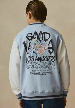 Person iført en lyseblå og hvid jakke med blomsterbroderi og tekst, der lyder ""Good Vibes Los Angeles California"" på ryggen.