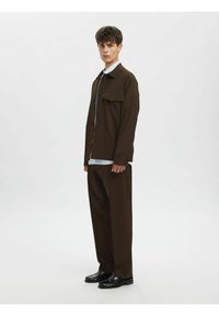 Brun jakke med en enkelt brystlomme, lagd over en lyseblå, stripete skjorte; kombinert med matchende brune bukser og svarte sko.