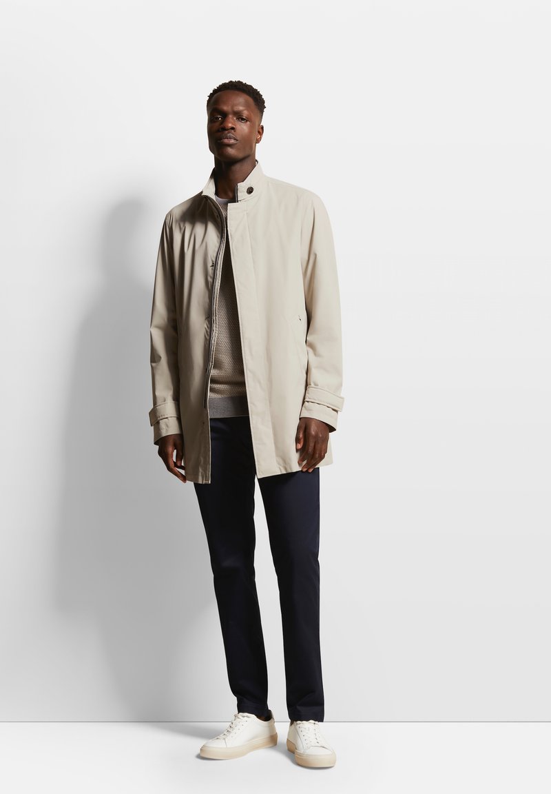 Veste courte beige avec un col haut, fermeture éclair et poches latérales, portée sur un pull léger et associée à un pantalon marine et des baskets blanches.