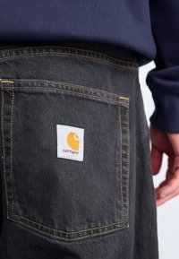 Primer plano de una persona que lleva pantalones de mezclilla negros de Carhartt con una etiqueta de logo amarilla en el bolsillo trasero y un sudadera azul marino.