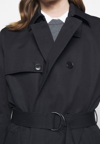 Femme portant un trench-coat noir ceinturé sur un pull gris et une chemise blanche à col, visible des épaules à la taille.