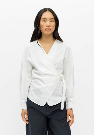 Femme aux longs cheveux foncés portant un chemisier blanc croisé noué à la taille et un pantalon noir, debout devant un fond blanc uni.