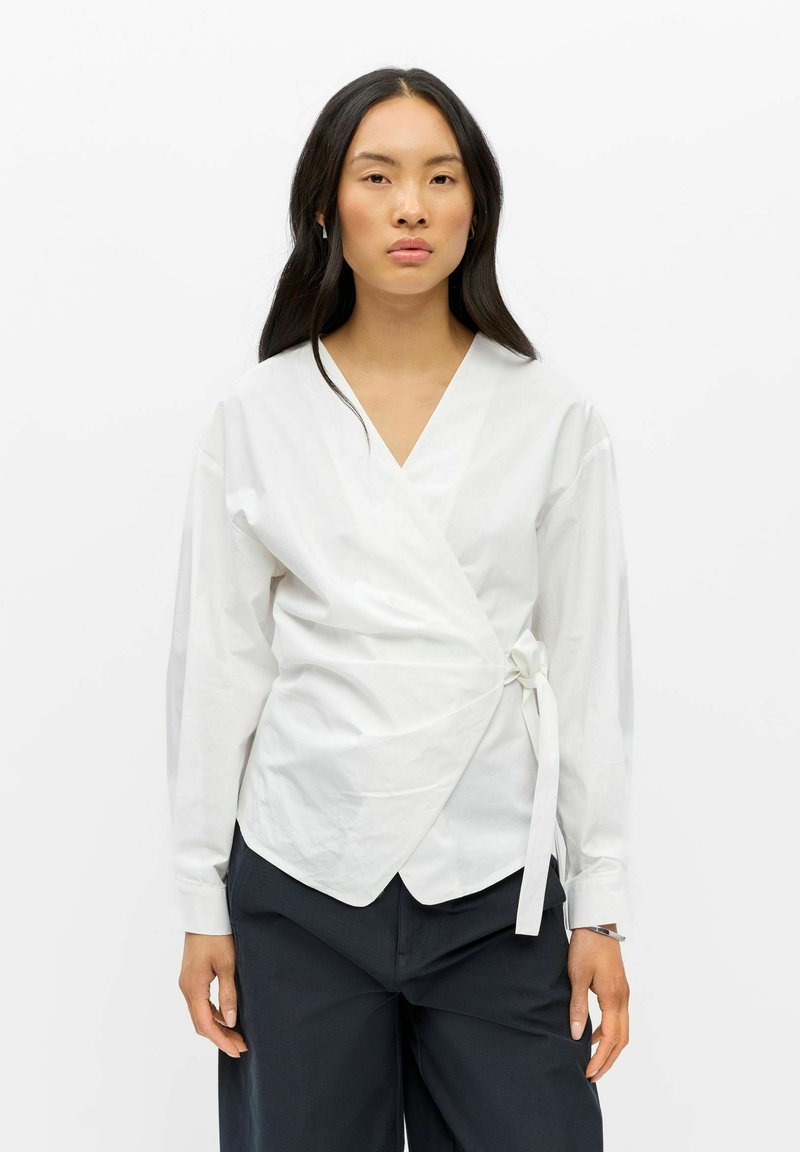 Femme aux longs cheveux foncés portant un chemisier blanc croisé noué à la taille et un pantalon noir, debout devant un fond blanc uni.