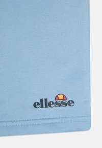 Tissu bleu clair avec une texture lisse, portant un logo noir "ellesse" et un graphique circulaire en orange et jaune en bas.