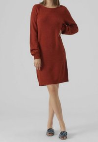 Robe-pull couleur rouille avec un col rond, des manches longues et une coupe décontractée. Texture douce et ourlet arrivant au genou. Portée avec des chaussures bleues à motifs floraux.