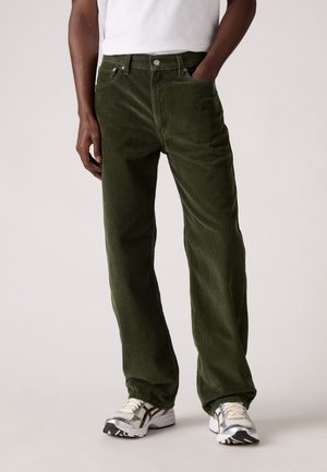 Man wearing olive green corduroy pants, white t-shirt, and white and silver sneakers, standing with one hand in pocket.
