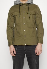 Hombre que lleva una chaqueta de botones verde oliva con bolsillos en el pecho y capucha gris, combinada con unos jeans negros desgastados y un accesorio de cadena.