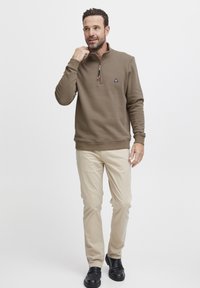 Felpa marrone con zip a quarantotto, tessuto morbido, polsini e orlo a coste, piccolo logo sul petto. Abbinata a pantaloni beige e scarpe nere.