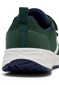 Nahaufnahme der Ferse eines grünen Sportschuhs, mit einer strukturierten Oberseite, einem Zugtab und einer weißen Sohle mit einer gemusterten blauen Außensohle.