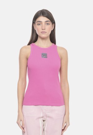 Donna con capelli castani lunghi che indossa un top smanicato rosa acceso a coste e pantaloni rosa chiaro, in piedi davanti a uno sfondo bianco semplice.