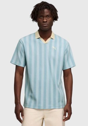 Hombre de piel oscura y rastas cortas vestido con una camiseta polo de rayas verticales azul claro con cuello beige y pantalones crema, de pie.