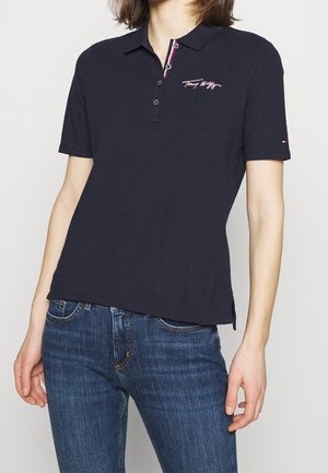 Vrouw draagt een marineblauw Tommy Hilfiger poloshirt met korte mouwen en een blauwe spijkerbroek, tegen een effen witte achtergrond.