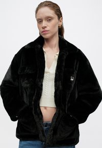 Chaqueta de piel sintética negra con una textura suave, gran cuello y dos bolsillos frontales; llevada sobre un top corto de punto trenzado color crema.