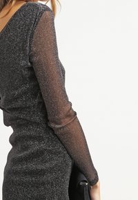 Musta mekko, jossa on kimaltava pinta, pitkät l äpinäkyvät hihat ja syvä V-mallinen selkäosa, lisänä musta käsilaukku.