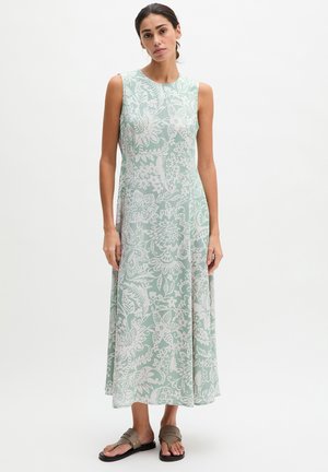 Kobieta w jasnozielonej, bezrękawnikowej sukience midi z białym kwiatowym wzorem i brązowych płaskich sandałach, stojąca na tle jednolitej białej ściany.