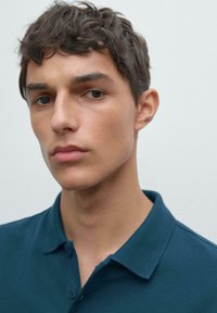Mladý muž s krátkými kudrnatými vlasy, oblečený v tmavě modré košili s límečkem, dívající se přímo do kamery, na jednobarevném světlém pozadí.