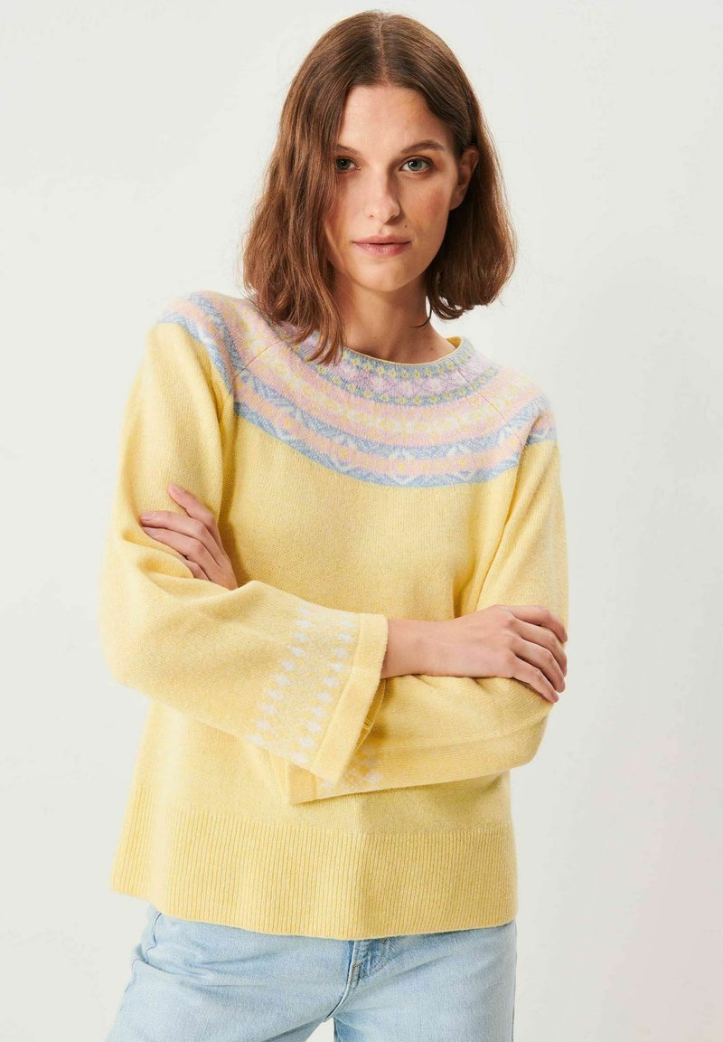 Kvinde med brunt hår iført en gul strikket sweater med pastelfarvede mønstre på bærestykket og lyseblå jeans, armene krydset.